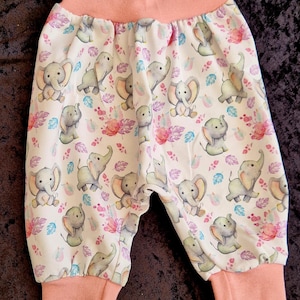 Könnte beinhalten: Babyhose mit weißem Hintergrund und einem sich wiederholenden Muster aus grauen Elefanten sowie rosafarbenen, violetten und türkisfarbenen Blättern. Das Kleidungsstück hat einen breiten, pfirsichfarbenen elastischen Bund und passende Beinbündchen.