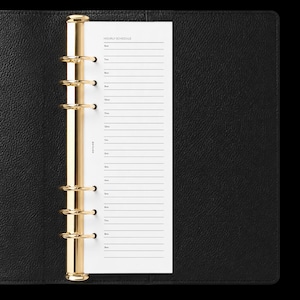May include: A black leather planner featuring a gold-toned six-ring metal binder mechanism. The white paper insert displays an hourly schedule with lines from 6am to 9pm and the text NOTAIRE printed vertically along the left margin of the page.