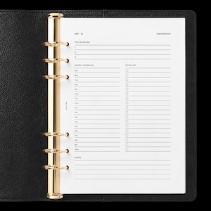 May include: A black leather planner with a gold-toned six-ring metal binder mechanism. The white paper insert features a daily schedule layout with sections for top priorities, an hourly schedule from 5am to 10pm, a to-do list, and notes.