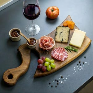 Puede incluir: Una tabla de charcutería de madera con varios quesos, embutidos, uvas y albaricoques secos. También hay una copa de vino tinto, pequeños cuencos de mermelada y un tomate. La tabla tiene un asa única y está colocada sobre una superficie oscura.