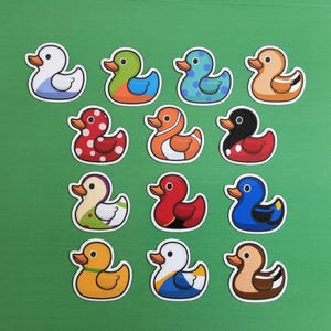 May include: Twelve duck stickers in various colours and patterns, arranged on a green surface. The ducks feature designs such as polka dots, stripes, and solid colours. Each duck has a unique colour scheme, including red, blue, yellow, and orange.