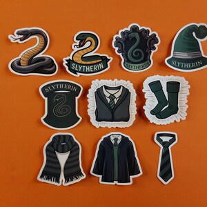 May include: Nine stickers featuring the Hogwarts house Slytherin. The stickers feature a snake, a crest, a hat, a scarf, a tie, a robe, and a pair of socks. All stickers have the word "Slytherin" on them.
