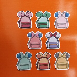May include: Eight colourful backpack stickers, each with a cartoon design featuring a bow and mouse ears. The backpacks are in various colours, including pink, green, blue, and yellow, set against an orange background.