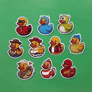 May include: A collection of ten vinyl stickers featuring yellow rubber ducks dressed as various animated characters. The designs include ducks in superhero, monster, cowboy, explorer, and racing-themed outfits with vibrant colours and white borders.
