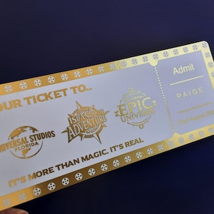 Pu&ograve; includere: Un biglietto dorato con testo bianco che recita "Your Ticket To..." e presenta i loghi di Universal Studios Florida, Islands of Adventure ed Epic Universe. Il biglietto &egrave; per Paige ed &egrave; datato 20 agosto 2005.