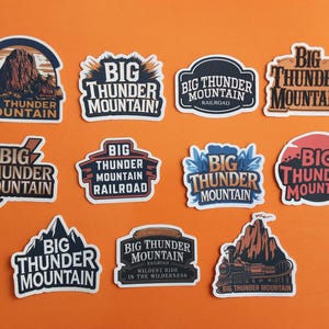 May include: A collection of Big Thunder Mountain stickers with various designs. Some stickers feature the text "Big Thunder Mountain" with mountain illustrations, while others include "Big Thunder Mountain Railroad". The stickers are on an orange background.