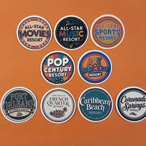 May include: Nine colourful round stickers with text and images. The stickers feature the names of different Disney resorts, including All-Star Movies, All-Star Music, All-Star Sports, Pop Century, Art of Animation, Port Orleans French Quarter, Caribbean Beach, and Coronado Springs.