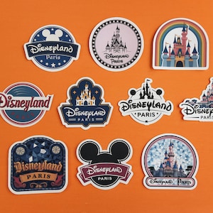 May include: A selection of Disneyland Paris stickers in various shapes and designs. The stickers feature the Disneyland logo, castle illustrations, and the text "Disneyland Paris" in different fonts and colours. The background is orange.