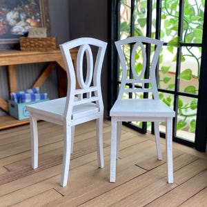 May include: Two white wooden chairs with an oval-shaped cutout in the back. The chairs have a simple design with straight legs and a rectangular seat.
