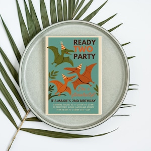 May include: A teal birthday invitation with orange and green pterodactyl illustrations. The text reads "READY TWO PARTY" and "IT'S MAXIE'S 2ND BIRTHDAY" with event details. The invitation is on a gray plate with a palm leaf.