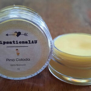 Puede incluir: Un pequeño tarro transparente con bálsamo labial amarillo en su interior. El tarro tiene una etiqueta que dice "LipsationalAU" y "Pina Colada" con un pequeño corazón y una flecha.