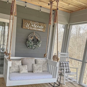 May include: White porch swing with rope hanging from the ceiling. The swing has a white cushion and pillows with a floral pattern. A wooden sign above the swing reads "Welcome to our porch".