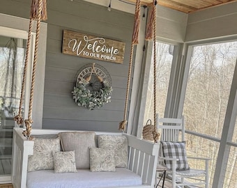 May include: White porch swing with rope hanging from the ceiling. The swing has a white cushion and pillows with a floral pattern. A wooden sign above the swing reads "Welcome to our porch".