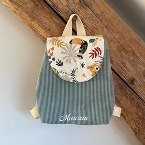 May include: A sage green corduroy backpack featuring a rounded flap with a colorful jungle animal print including a toucan and sloth. The name Marceau is embroidered in white script on the front. The bag has beige webbing straps and a top handle.