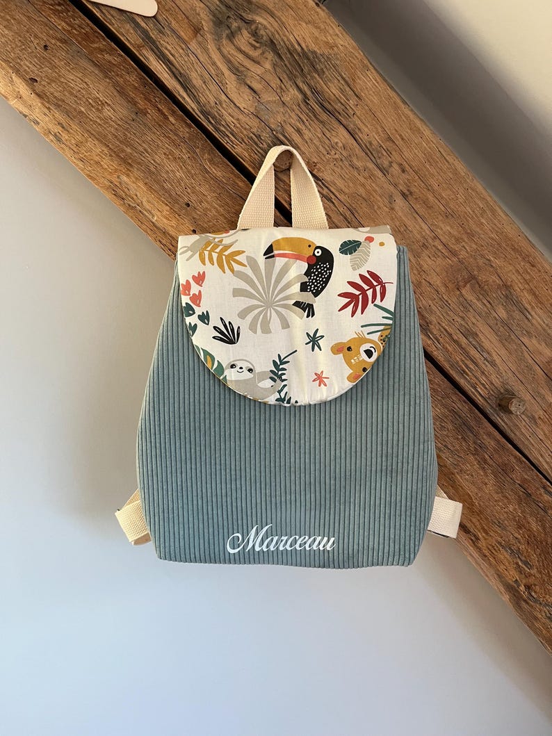 May include: A sage green corduroy backpack featuring a rounded flap with a colorful jungle animal print including a toucan and sloth. The name Marceau is embroidered in white script on the front. The bag has beige webbing straps and a top handle.