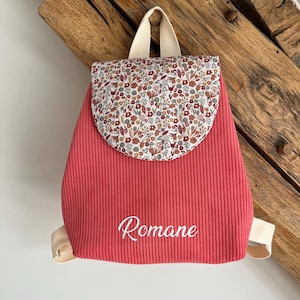 May include: A coral-colored corduroy backpack featuring a rounded flap with a multi-colored floral pattern in shades of red, brown, and grey. The front of the bag is embroidered with the name Romane in white script and includes beige straps.