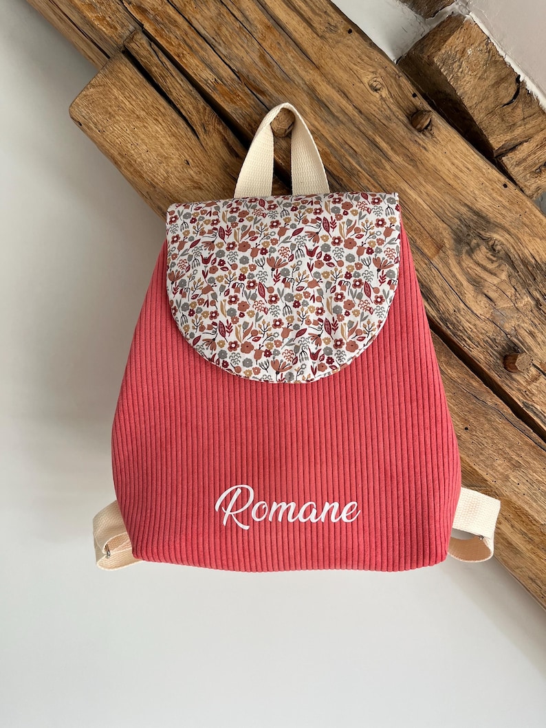 May include: A coral-colored corduroy backpack featuring a rounded flap with a multi-colored floral pattern in shades of red, brown, and grey. The front of the bag is embroidered with the name Romane in white script and includes beige straps.