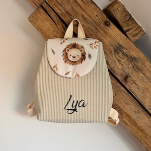 May include: A beige corduroy backpack featuring a flap with a printed lion face and leaf pattern. The name Lya is embroidered in black script on the front. The bag includes a top handle and adjustable shoulder straps in a matching beige fabric.
