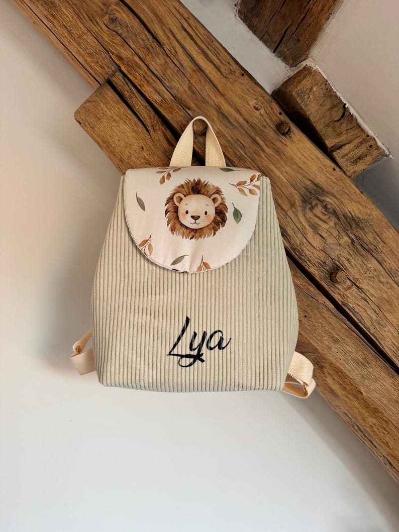May include: A beige corduroy backpack featuring a flap with a printed lion face and leaf pattern. The name Lya is embroidered in black script on the front. The bag includes a top handle and adjustable shoulder straps in a matching beige fabric.