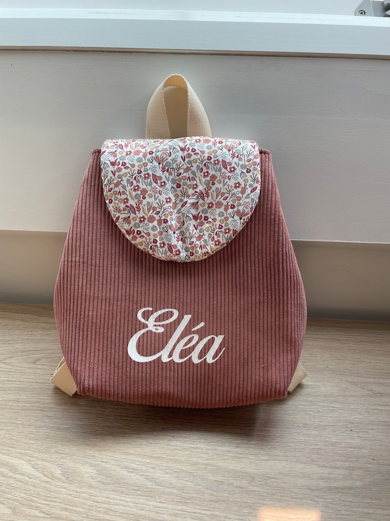 May include: A dusty rose corduroy backpack featuring a rounded flap with a multi-colored floral pattern. The name El&eacute;a is printed in white script on the front. The bag includes beige fabric straps and a top handle for carrying or hanging.
