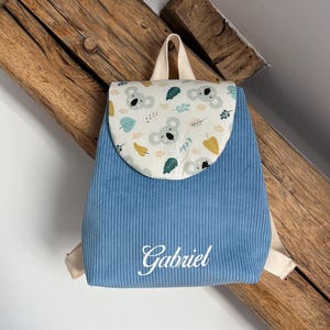May include: A blue corduroy backpack featuring a flap with a koala and leaf pattern in shades of gray, green, and yellow. The name Gabriel is embroidered in white script on the front. The bag includes beige fabric straps and a top handle.