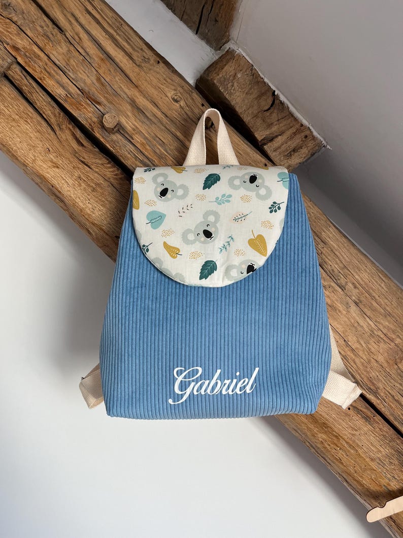 May include: A blue corduroy backpack featuring a flap with a koala and leaf pattern in shades of gray, green, and yellow. The name Gabriel is embroidered in white script on the front. The bag includes beige fabric straps and a top handle.
