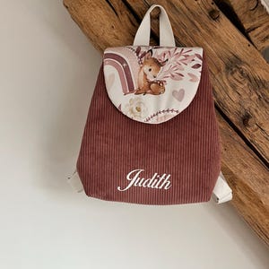 May include: A dusty rose corduroy backpack featuring a flap with a printed illustration of a fawn, a rainbow, and floral elements. The name Judith is embroidered in white script on the front pocket. The backpack has white straps and a top handle.