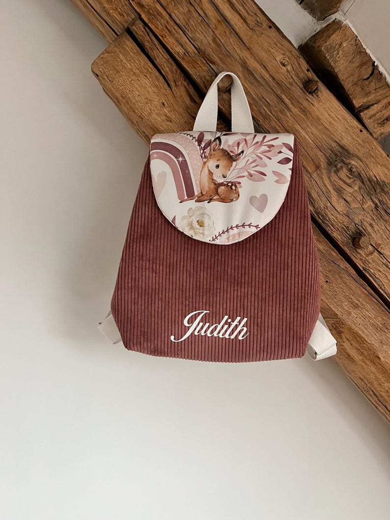 May include: A dusty rose corduroy backpack featuring a flap with a printed illustration of a fawn, a rainbow, and floral elements. The name Judith is embroidered in white script on the front pocket. The backpack has white straps and a top handle.