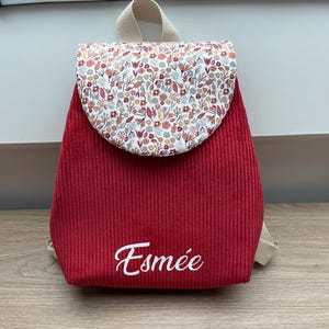 May include: A red corduroy backpack featuring a rounded flap with a multi-colored floral pattern in shades of pink, orange, and green. The name Esm&eacute;e is embroidered in white script on the lower front section, and the bag has beige straps.
