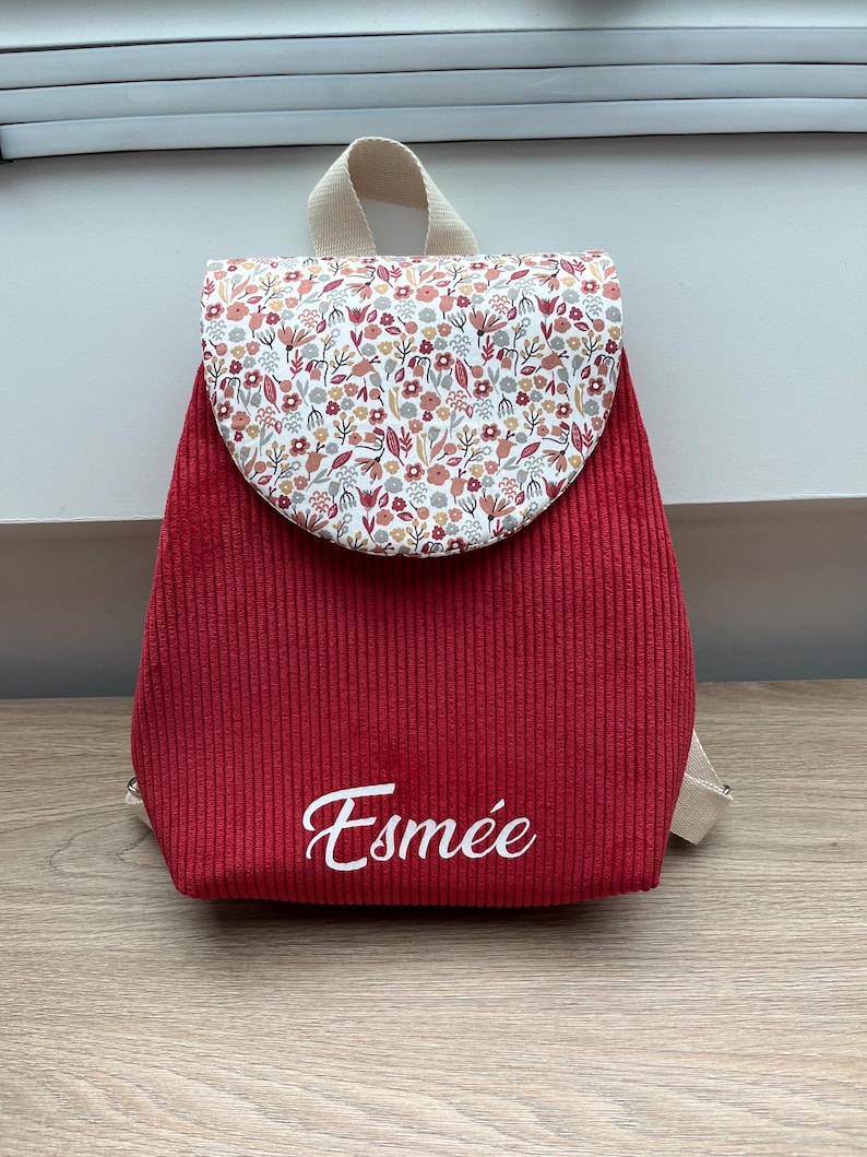 May include: A red corduroy backpack featuring a rounded flap with a multi-colored floral pattern in shades of pink, orange, and green. The name Esm&eacute;e is embroidered in white script on the lower front section, and the bag has beige straps.