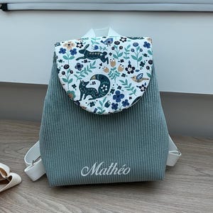 May include: A sage green corduroy backpack featuring a white flap with a colorful woodland print of foxes, rabbits, birds, and flowers. The name Math&eacute;o is embroidered in white script on the front. The bag includes white adjustable shoulder straps.