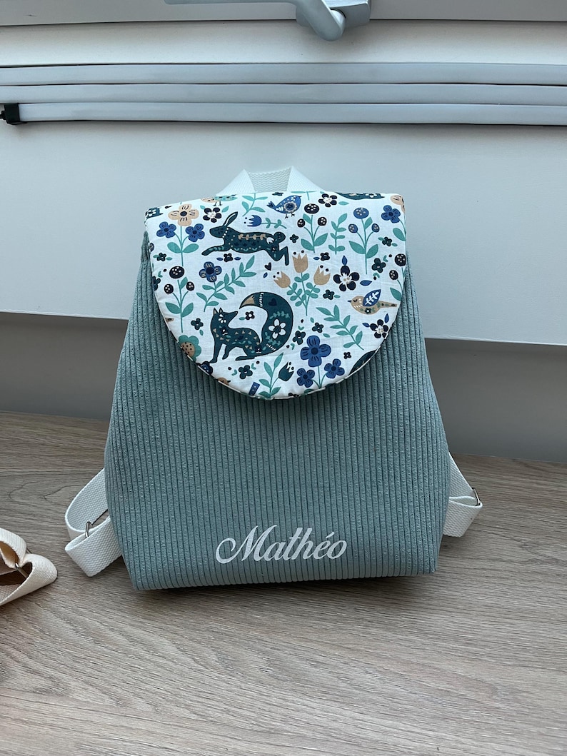 May include: A sage green corduroy backpack featuring a white flap with a colorful woodland print of foxes, rabbits, birds, and flowers. The name Math&eacute;o is embroidered in white script on the front. The bag includes white adjustable shoulder straps.