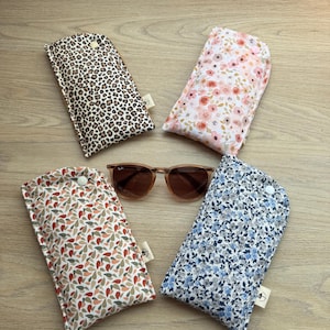 Peut inclure: Quatre étuis à lunettes rectangulaires en tissu avec fermetures à pression, présentant des motifs léopard, fleurs roses, feuilles multicolores et fleurs bleues, disposés autour d'une paire de lunettes de soleil à verres bruns.