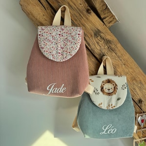 May include: Two corduroy backpacks with top handles and flap closures. The left backpack is dusty pink with a floral print flap and the name Jade embroidered in white. The right backpack is sage green with a lion print flap and the name L&eacute;o embroidered in white.