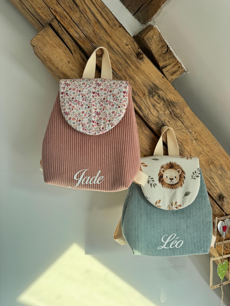 May include: Two corduroy backpacks with top handles and flap closures. The left backpack is dusty pink with a floral print flap and the name Jade embroidered in white. The right backpack is sage green with a lion print flap and the name L&eacute;o embroidered in white.