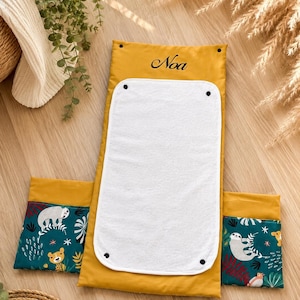 Peut inclure: Un matelas à langer rectangulaire jaune moutarde avec le prénom Noa brodé en écriture noire. Le centre comporte un tapis en tissu blanc amovible fixé par des boutons-pression noirs. Deux panneaux latéraux assortis affichent un motif jungle.
