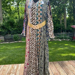May include: A long, patchwork dress with a V-neck and long sleeves. The dress features a variety of patterns and colors, including floral, paisley, and geometric designs. A gold-colored belt cinches the waist. The dress is displayed on a black mannequin.