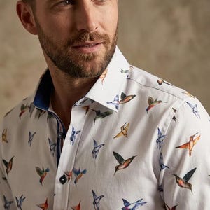 Peut inclure: Chemise blanche à motifs de colibris colorés. La chemise a un col classique et une bordure bleue à l'intérieur du col et des poignets. Les colibris sont représentés dans diverses couleurs, dont le bleu, l'orange et le vert.