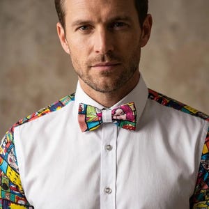 Op de afbeelding: Een wit overhemd met knoopsluiting en een kleurrijke stripboekprint op de mouwen en schouders, gecombineerd met een bijpassende vlinderdas. Het levendige grafische patroon bevat diverse pop-art illustraties, tekstballonnen en primaire kleuren.