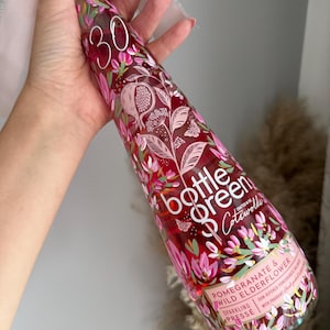 May include: A clear glass bottle of Bottle Green Pomegranate & Wild Elderflower sparkling pressé. The bottle features a vibrant floral and pomegranate graphic print in shades of pink, red, and green, with the text "30" printed near the neck.