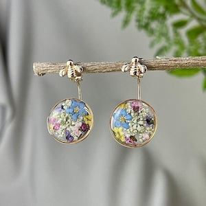 Può includere: Un paio di orecchini dorati con un design a forma di ape. Ogni orecchino presenta un pendente rotondo trasparente riempito di fiori secchi colorati. I fiori includono petali blu, rosa e bianchi, creando un aspetto delicato e naturale.