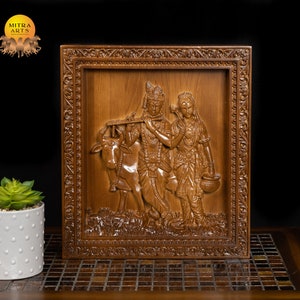 K&ouml;nnte beinhalten: Eine Holzschnitzerei f&uuml;r die Wand, die Radha Krishna, eine hinduistische Gottheit, darstellt, die Fl&ouml;te spielt, mit einer Kuh an seiner Seite. Die Schnitzerei ist in einem dunklen Holzrahmen mit aufwendigen Details gerahmt.