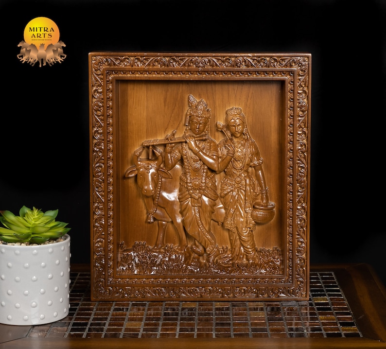 K&ouml;nnte beinhalten: Eine Holzschnitzerei f&uuml;r die Wand, die Radha Krishna, eine hinduistische Gottheit, darstellt, die Fl&ouml;te spielt, mit einer Kuh an seiner Seite. Die Schnitzerei ist in einem dunklen Holzrahmen mit aufwendigen Details gerahmt.