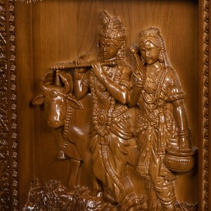 K&ouml;nnte beinhalten: Eine Holzschnitzerei, die ein Paar, m&ouml;glicherweise Radha und Krishna, mit einer Kuh zeigt. Die Schnitzerei ist detailliert und kunstvoll, die Figuren tragen aufwendige Kleidung und Schmuck. Die Schnitzerei ist in einem Holzrahmen mit verzierten Details gefasst.