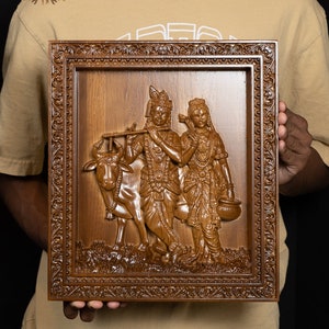 K&ouml;nnte beinhalten: Ein Wandbild aus Holz, das eine geschnitzte Szene von Krishna und Radha mit einer Kuh darstellt. Das Kunstwerk ist in einem dunkelbraunen Holzrahmen mit aufwendigen Schnitzereien gerahmt. Der Rahmen hat einen erh&ouml;hten Rand und einen dekorativen Rand.