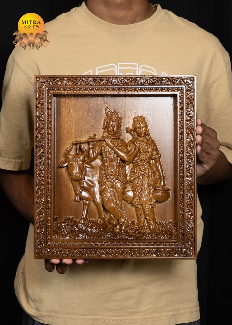 K&ouml;nnte beinhalten: Ein Wandbild aus Holz, das eine geschnitzte Szene von Krishna und Radha mit einer Kuh darstellt. Das Kunstwerk ist in einem dunkelbraunen Holzrahmen mit aufwendigen Schnitzereien gerahmt. Der Rahmen hat einen erh&ouml;hten Rand und einen dekorativen Rand.