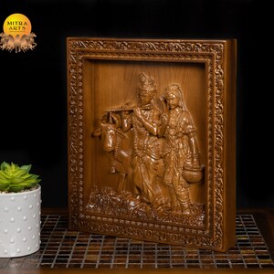 K&ouml;nnte beinhalten: Eine Holzschnitzerei f&uuml;r die Wand, die Radha Krishna, ein hinduistisches G&ouml;tterpaar, mit einer Kuh darstellt. Die Schnitzerei ist in einem dunklen Holzrahmen mit aufwendigen Details gerahmt.