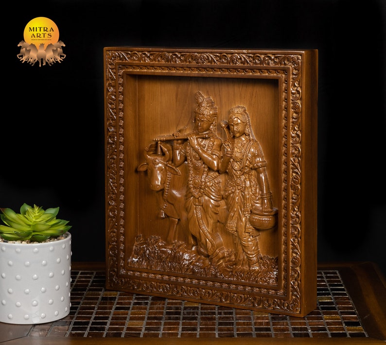 K&ouml;nnte beinhalten: Eine Holzschnitzerei f&uuml;r die Wand, die Radha Krishna, ein hinduistisches G&ouml;tterpaar, mit einer Kuh darstellt. Die Schnitzerei ist in einem dunklen Holzrahmen mit aufwendigen Details gerahmt.