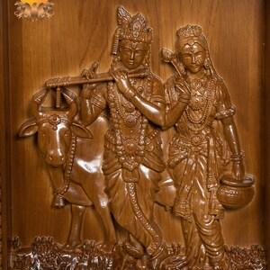 K&ouml;nnte beinhalten: Eine Holzschnitzerei von Krishna und Radha, wobei Krishna eine Fl&ouml;te spielt und Radha einen Topf h&auml;lt. Die Schnitzerei ist detailliert und kunstvoll, mit einer Kuh, die neben ihnen steht.
