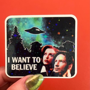 May include: A sticker featuring a holographic image of two people looking up at a UFO with the text "I WANT TO BELIEVE" in white letters.